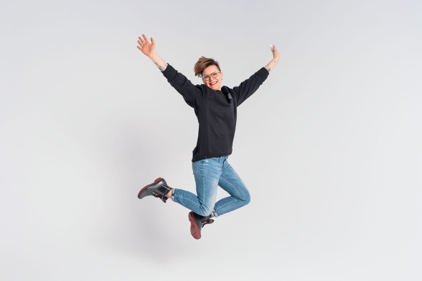 A person with short hair and glasses, wearing a black sweater and jeans, jumping joyfully against a plain white background. Their arms are outstretched and they have a big smile on their face, capturing a moment of excitement and happiness.