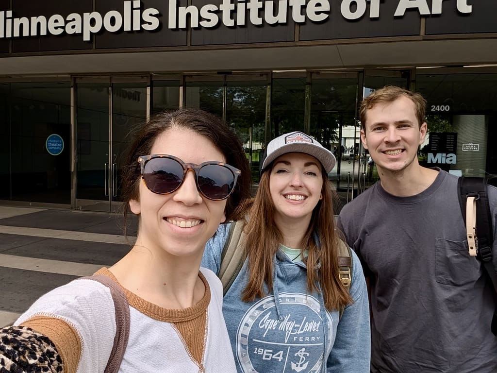 Three friends stand smiling in front of the entrance to the Minneapolis Institute of Art. The group includes a woman wearing large sunglasses and a white and brown top, a woman in a baseball cap and blue hoodie, and a man in a gray t-shirt. They are standing close together, with the sign for the Minneapolis Institute of Art clearly visible above the entrance. The background shows glass doors and part of the museum's interior. The atmosphere is friendly and cheerful, suggesting they are excited to visit the museum.