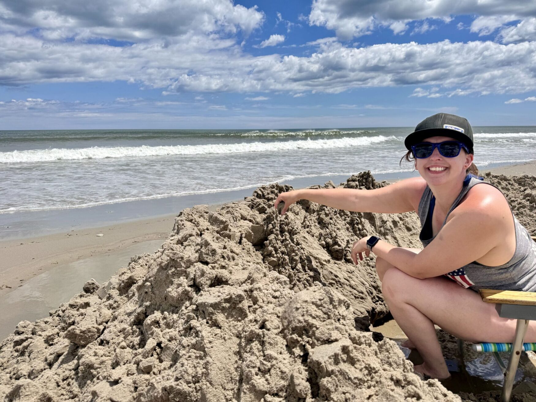 A woman wearing a black cap, sunglasses, and a gray tank top with an American flag design sits on a colorful beach chair by the ocean. She is smiling and building a large sandcastle with her hands. The beach is sandy with waves gently crashing on the shore. The sky is partly cloudy with patches of blue, indicating a sunny day. The atmosphere is relaxed and joyful, reflecting a pleasant day at the beach.