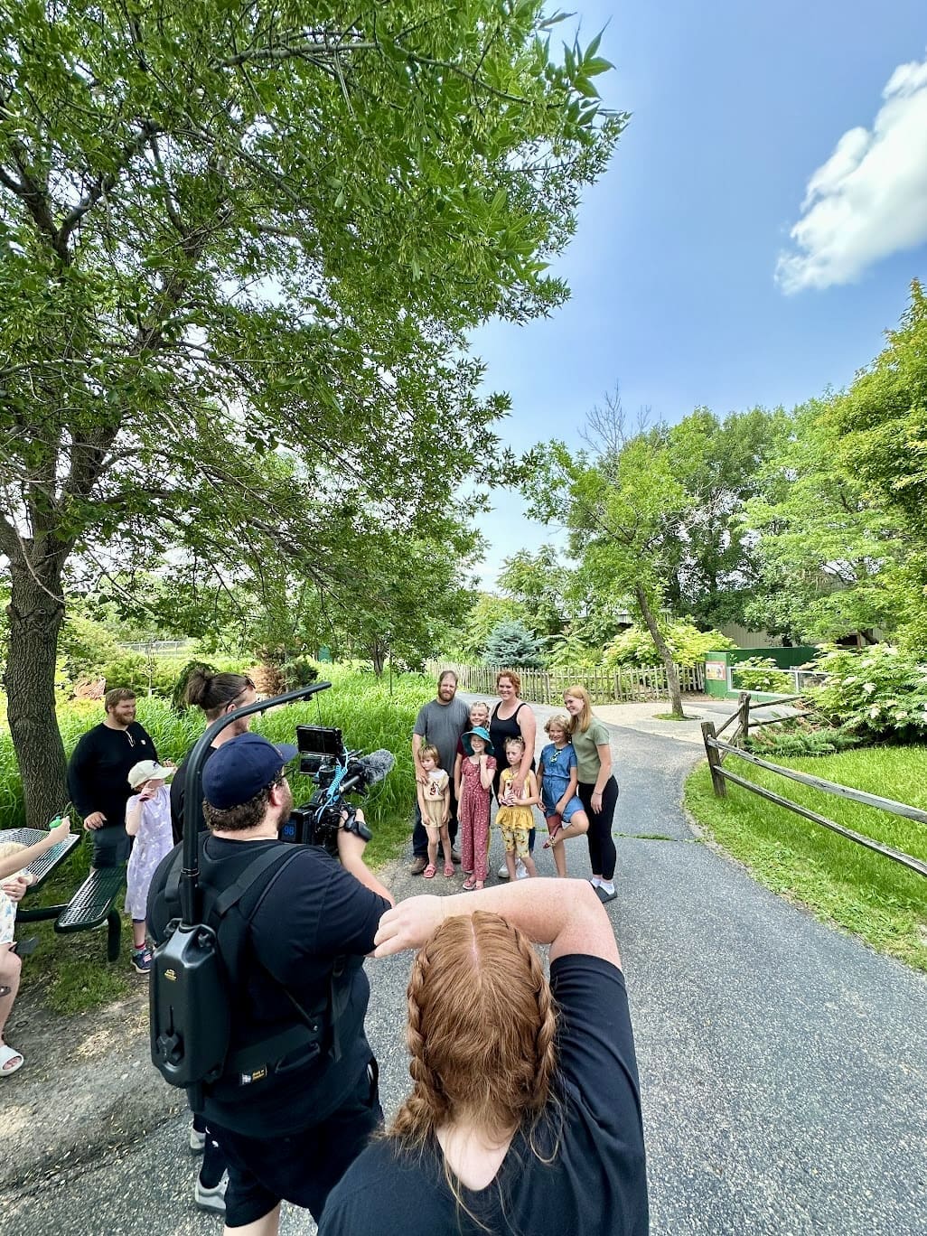 A camera crew films a group of adults and children standing together on a paved pathway in a park-like setting. The group is smiling and posing for the camera. The setting is lush and green, with tall trees providing shade. The pathway curves through the park, which features wooden fences, grassy areas, and various plants. The sky is clear and blue, indicating a sunny day. The camera crew is focused on capturing the group, with one cameraman holding a professional camera rig, and another person with red hair and braids standing nearby.