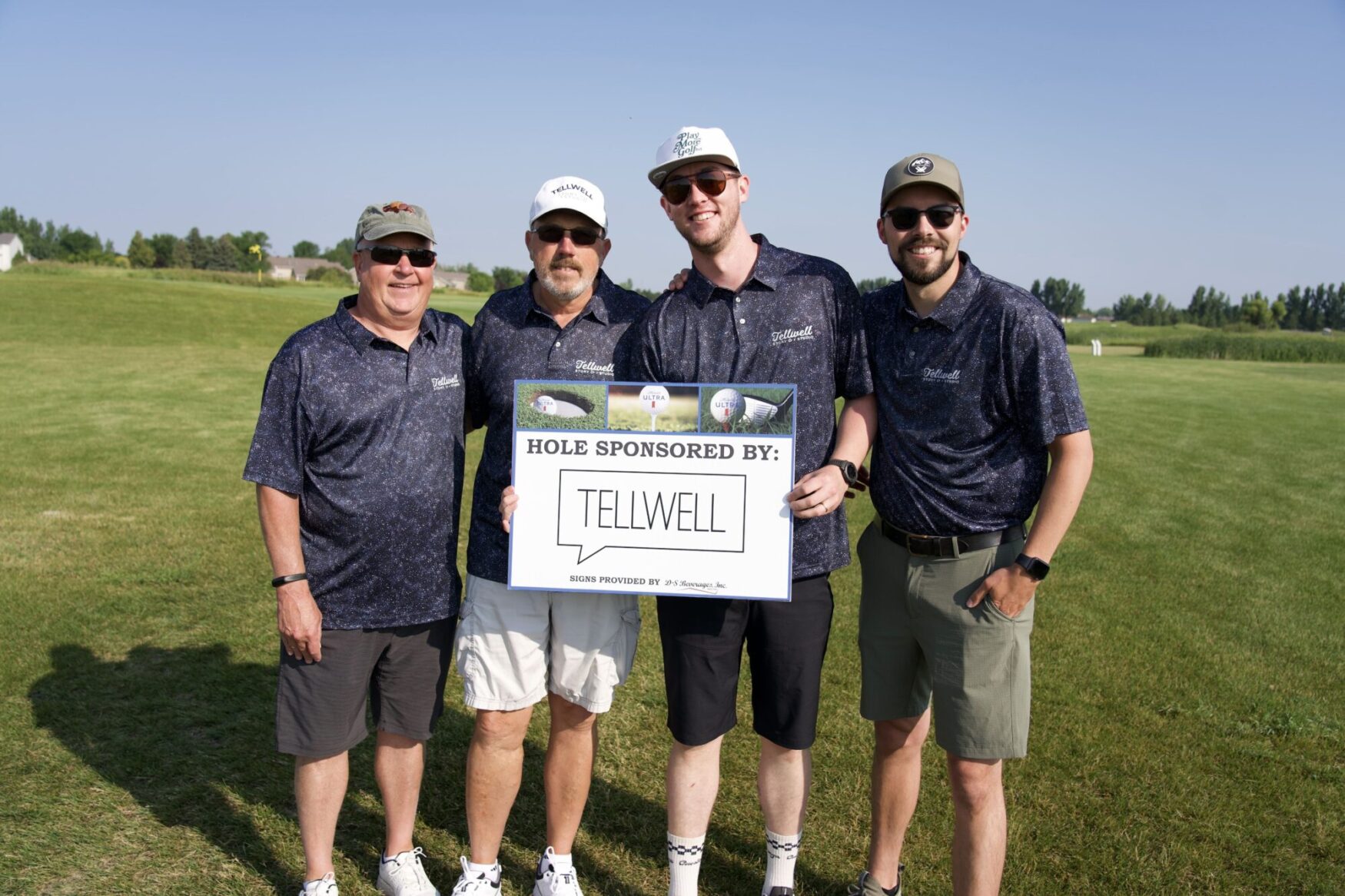 Four men stand on a golf course, smiling and posing with a sign that reads "Hole Sponsored By: Tellwell." They are all wearing matching Tellwell-branded polo shirts and sunglasses. The man on the left wears a baseball cap and the man second from the right wears a white hat with sunglasses. The group is standing on a sunny day with a clear blue sky and green grass in the background. The atmosphere is friendly and collaborative, indicating participation in a sponsored golf event.