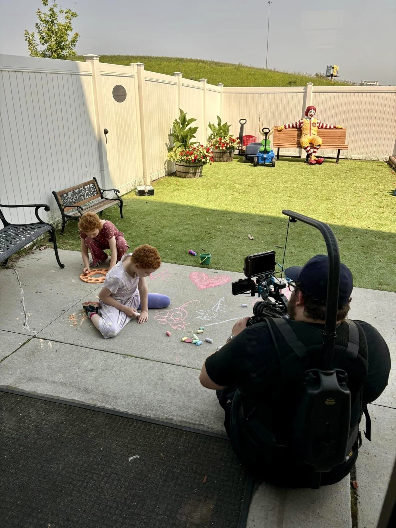 A camera operator films two young children drawing with colorful chalk on a concrete patio in a backyard. One child, wearing a purple outfit, is drawing a figure, while the other child, in a red outfit, watches. The patio is bordered by a white fence and features black metal benches and potted plants with flowers. In the background, a Ronald McDonald statue sits on a bench in the grassy area. The atmosphere is playful and creative, with various toys scattered around.