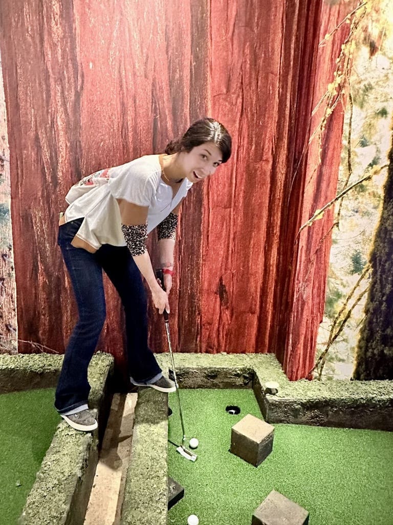 A woman with dark hair, wearing a white and brown top and blue jeans, is playing mini-golf indoors. She is standing on a narrow, elevated section of the course, holding a putter and preparing to hit the golf ball. The background features a mural of tall redwood trees, creating an outdoor forest atmosphere. The woman is smiling and appears to be enjoying the game. The mini-golf course has green artificial turf and small obstacles.