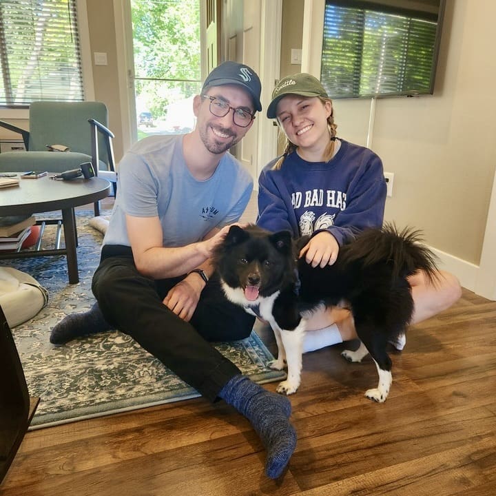 A young man and woman sit on the floor of a cozy living room, smiling at the camera. The man, wearing glasses, a light blue t-shirt, and a cap, sits cross-legged next to the woman, who is wearing a dark blue sweatshirt, shorts, and a cap. They both have their hands on a fluffy black and white dog that is standing between them, happily panting with its tongue out. The room has a soft rug, a coffee table with books, and a large window letting in natural light, creating a warm and inviting atmosphere.