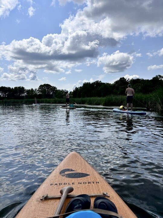 A scenic view from the perspective of a paddle boarder on a calm lake, showing the front of the board labeled "Paddle North." In the distance, three people are stand-up paddleboarding, with lush greenery and reeds lining the shore. The sky is partly cloudy, with sunlight filtering through, creating a serene and peaceful atmosphere. The water reflects the blue sky and clouds, enhancing the tranquility of the scene.