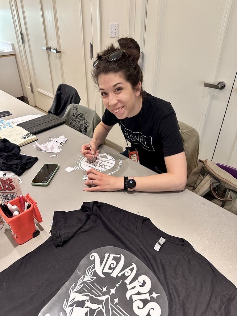A woman with dark hair tied in a bun, wearing a black "Tellwell" t-shirt and sunglasses on her head, sits at a table engaged in a craft project. She is using a tool to work on a design printed on a black t-shirt in front of her. The table is scattered with various materials, including a phone, markers, and other craft supplies. The woman is smiling and appears to be enjoying her activity. The setting is a bright indoor space with white walls and a door in the background.