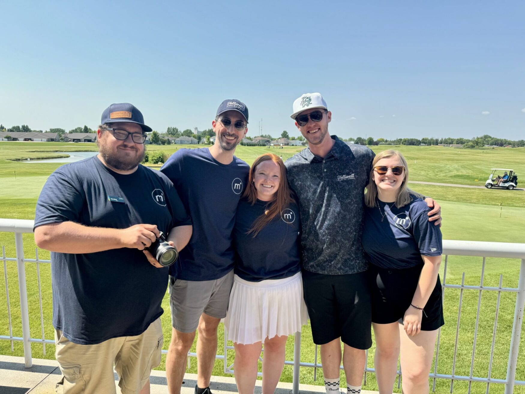 A group of five people stand together on a balcony overlooking a golf course on a sunny day. They are all smiling and wearing matching navy blue shirts with logos. The group includes three men and two women, with one man holding a camera. The golf course behind them features green fairways, a golf cart, and a water hazard, set against a clear blue sky. The setting appears to be a professional or corporate outing, with the group enjoying their time outdoors.