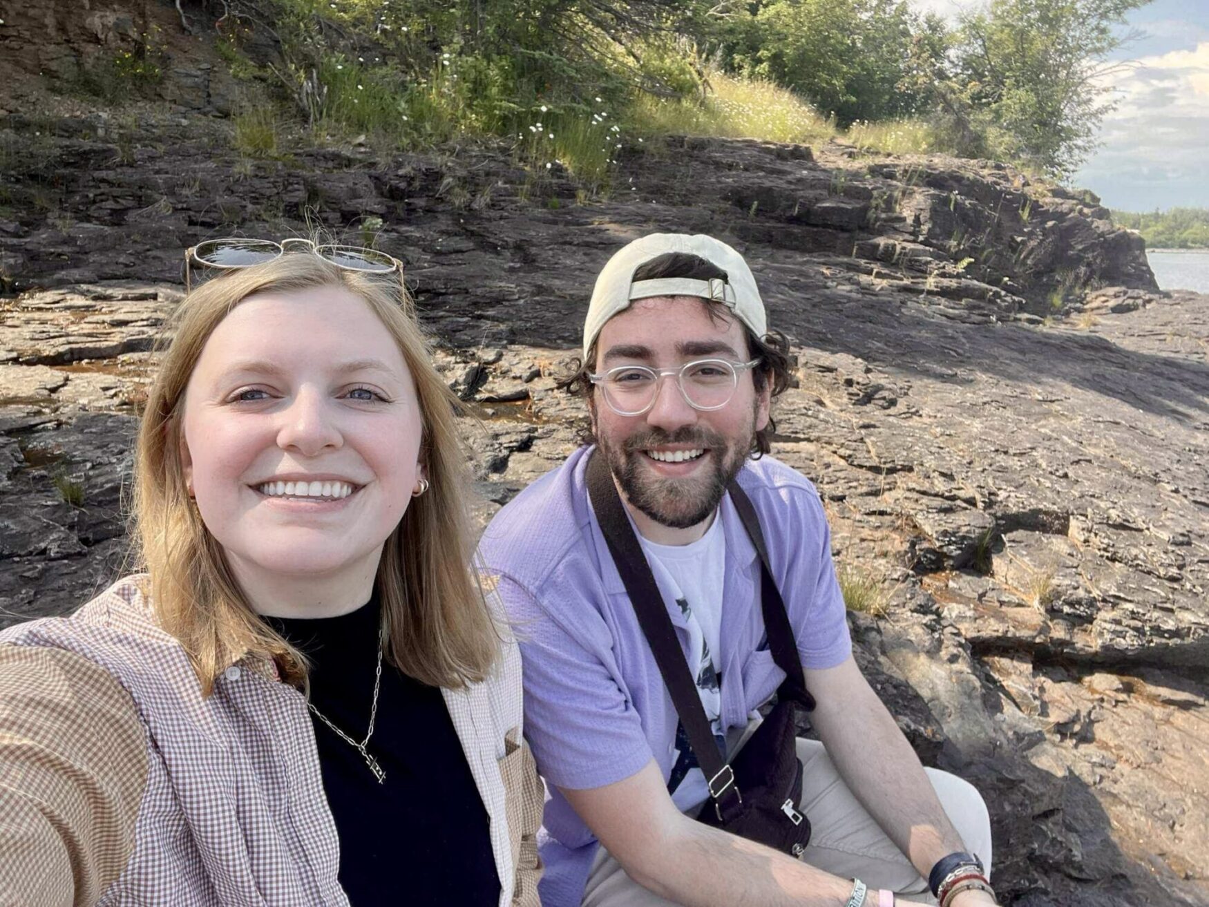 A young woman and man sit on rocky terrain, smiling at the camera. The woman, with shoulder-length blonde hair and wearing a checkered shirt over a black top, has sunglasses perched on her head. The man, with dark hair, a beard, glasses, and wearing a light purple shirt over a white tee, has a baseball cap worn backward. The background features rocky outcrops, sparse greenery, and a glimpse of water, indicating they are outdoors in a natural setting on a sunny day.