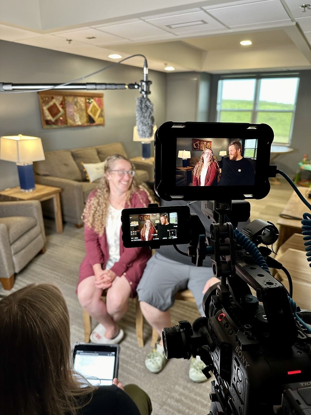 A behind-the-scenes view of an interview being conducted in a cozy, well-lit room. A woman with curly blonde hair and glasses, wearing a maroon cardigan, and a man with a beard, wearing a black shirt and gray shorts, are sitting on wooden chairs, smiling and looking at each other. A camera crew is filming them, with a professional camera and microphone visible in the foreground. The camera's display screen shows a close-up of the couple. In the background, there are comfortable chairs, a side table with a lamp, and a large window providing natural light.
