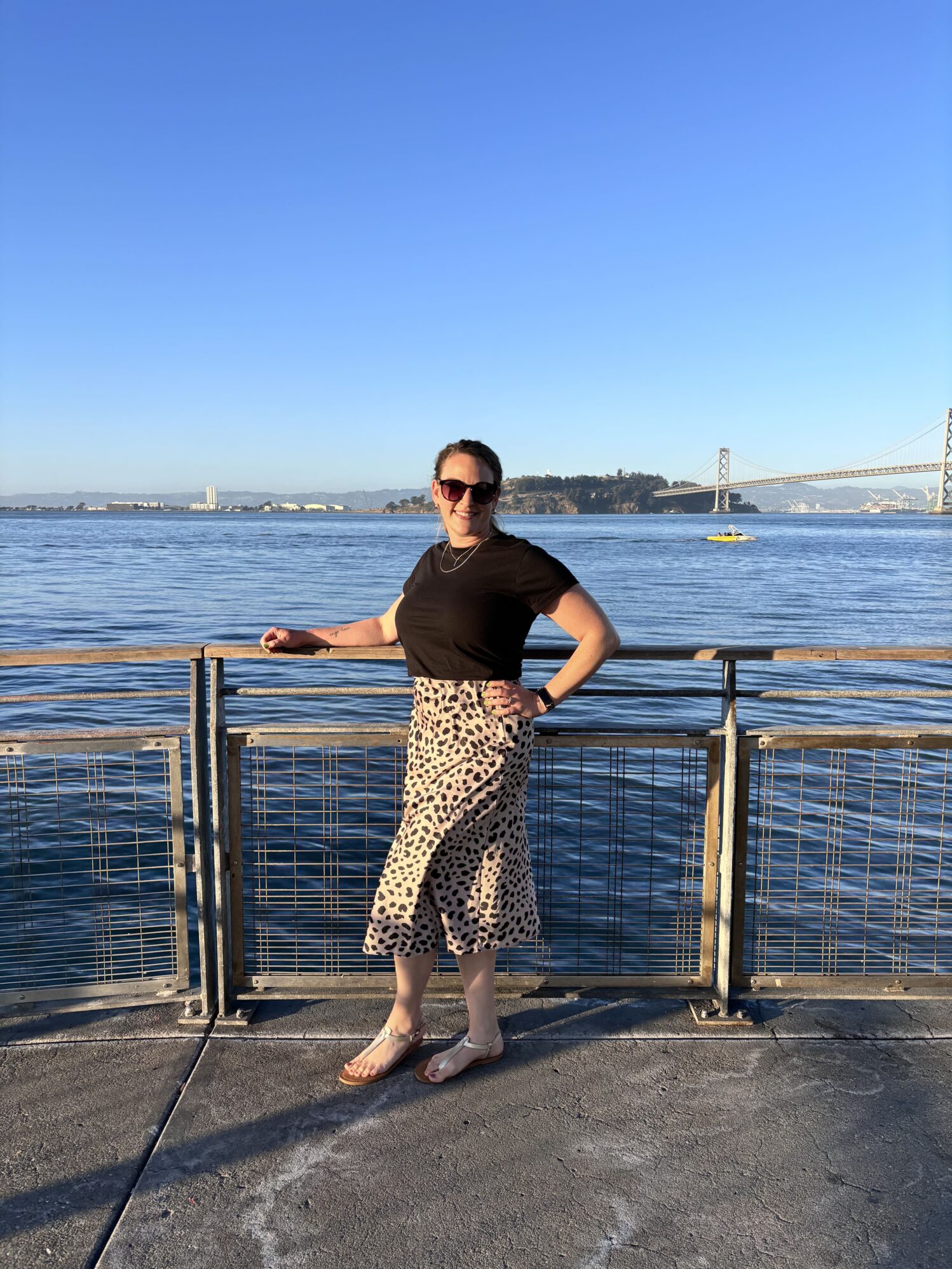 A woman wearing sunglasses and a black shirt stands smiling by the waterfront with the San Francisco Bay and a distant bridge in the background on a sunny day.