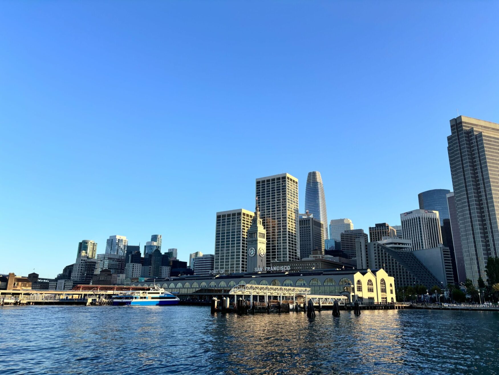 A view of San Francisco's skyline with the Ferry Building and waterfront in the foreground under a clear blue sky.