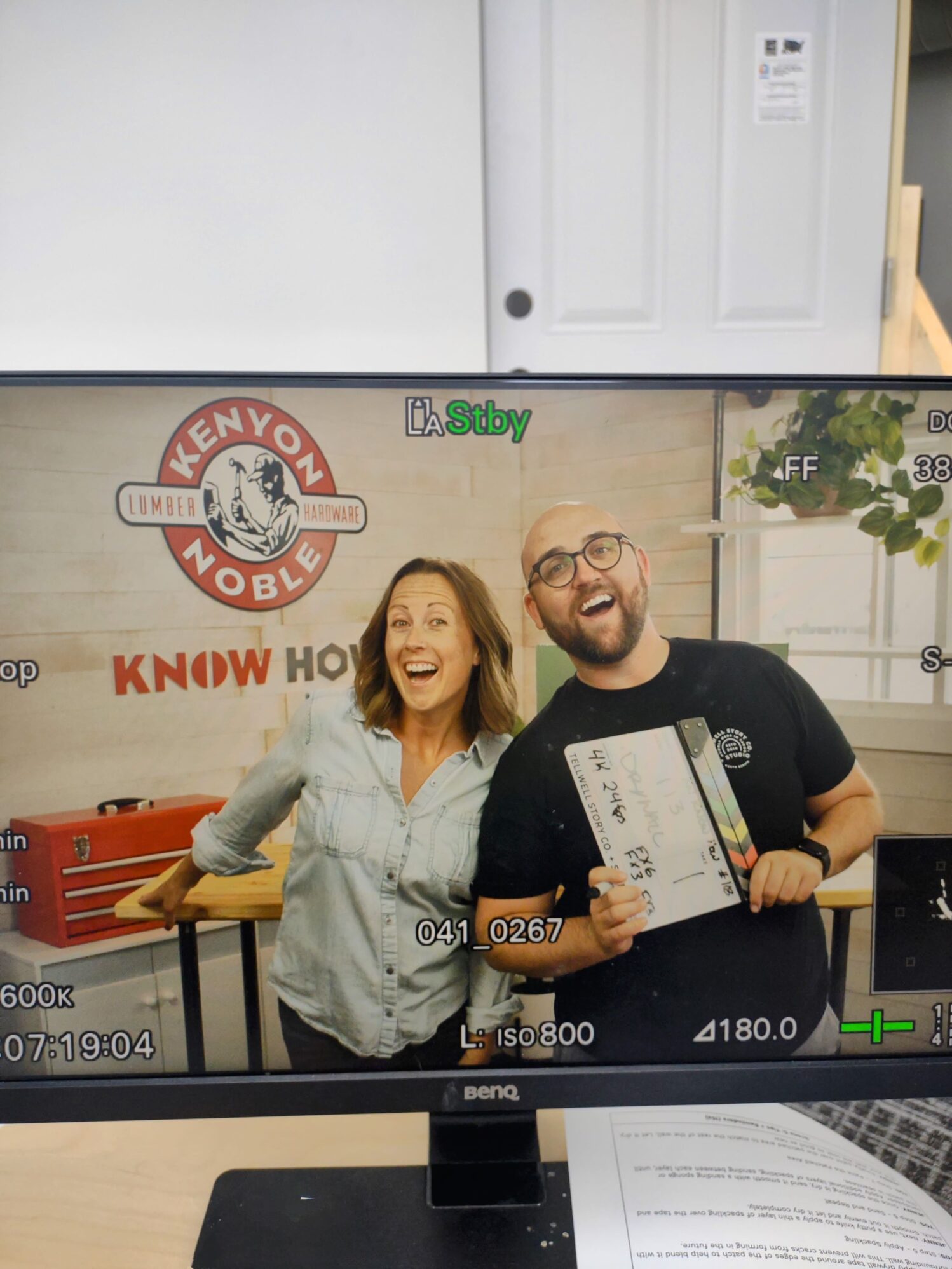 A monitor displaying a camera view of a woman and a man standing together and smiling in a studio. The woman, wearing a light denim shirt, is leaning on a table, while the man, wearing glasses and a black shirt, is holding a clapperboard. Behind them, a sign reads 'Kenyon Noble Lumber & Hardware' with 'Know How' below it. The monitor shows various camera settings and information around the edges of the screen.