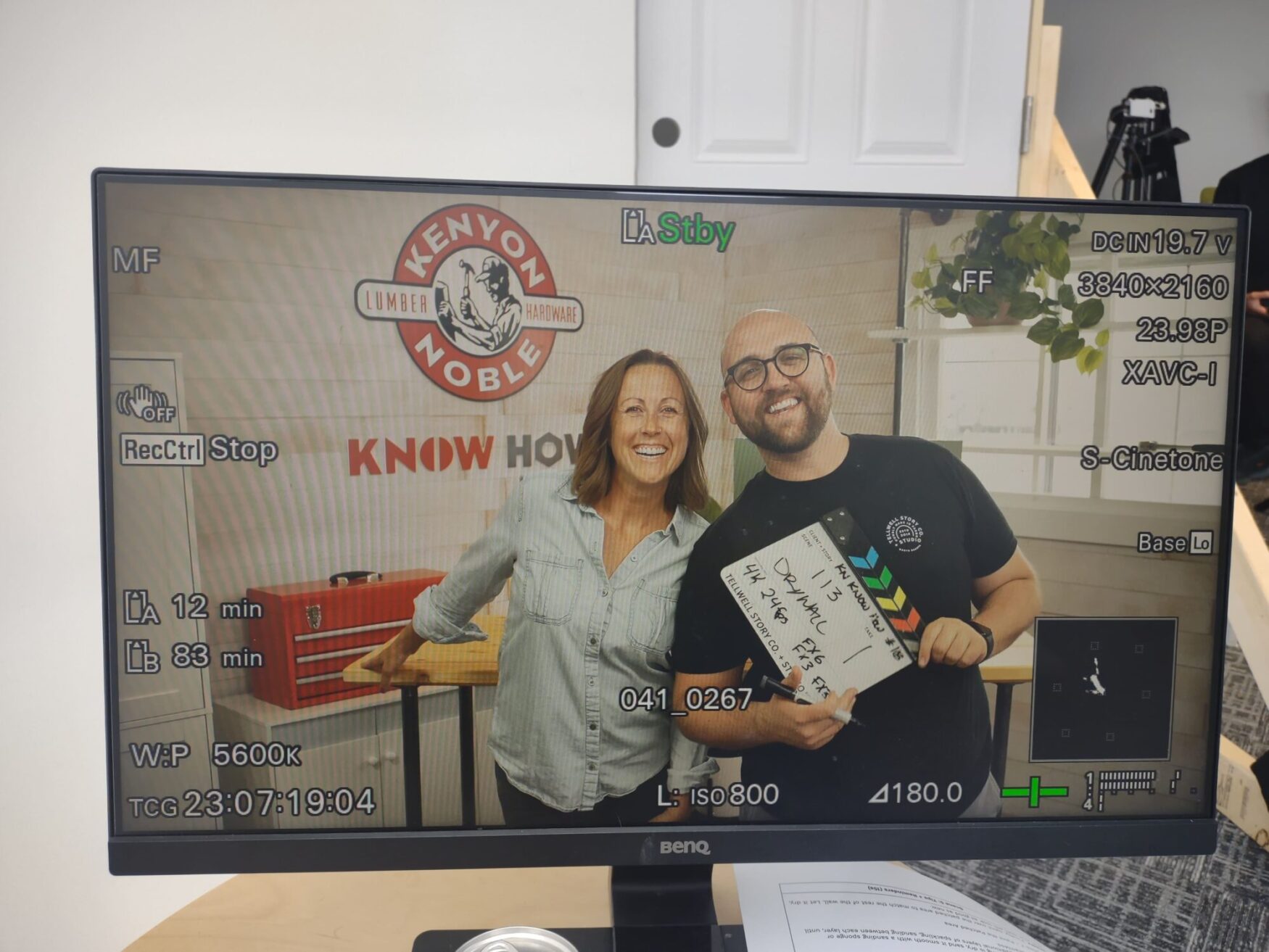 A monitor displaying a camera view of two smiling people standing side by side in a studio. The woman on the left is wearing a light denim shirt, and the man on the right, wearing glasses and a black shirt, is holding a clapperboard. Behind them, a sign reads 'Kenyon Noble' with the tagline 'Lumber & Hardware' and 'Know How' below it. The monitor shows various camera settings and information around the edges of the screen