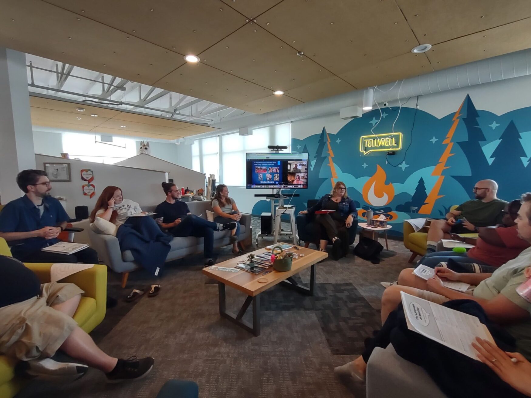 A group of people sitting in a casual meeting room with colorful, modern decor. They are seated on sofas and chairs arranged in a circle, with a coffee table in the center. A large screen on the far wall displays a video conference. The wall behind the screen features a mural with trees, a campfire, and the word 'Tellwell' in neon lights. The participants appear engaged in discussion, holding papers and notebooks.