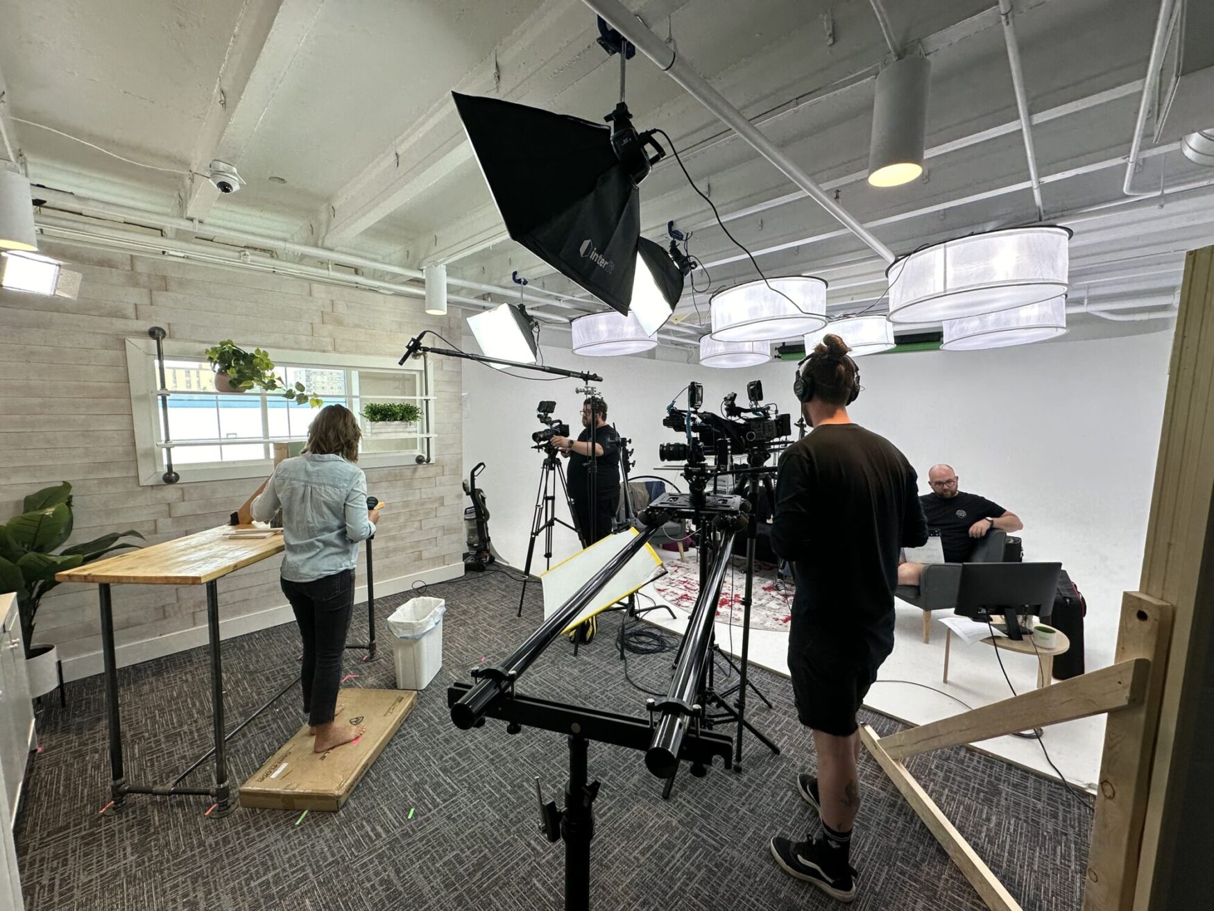 A behind-the-scenes view of a filming studio setup with multiple cameras, lights, and crew members. A woman is standing at a wooden table in the left foreground, while a camera operator with headphones stands nearby. In the background, another crew member is sitting on a couch with a laptop. The studio has a modern and organized setup with various equipment and plants decorating the space, and large lights hanging from the ceiling.