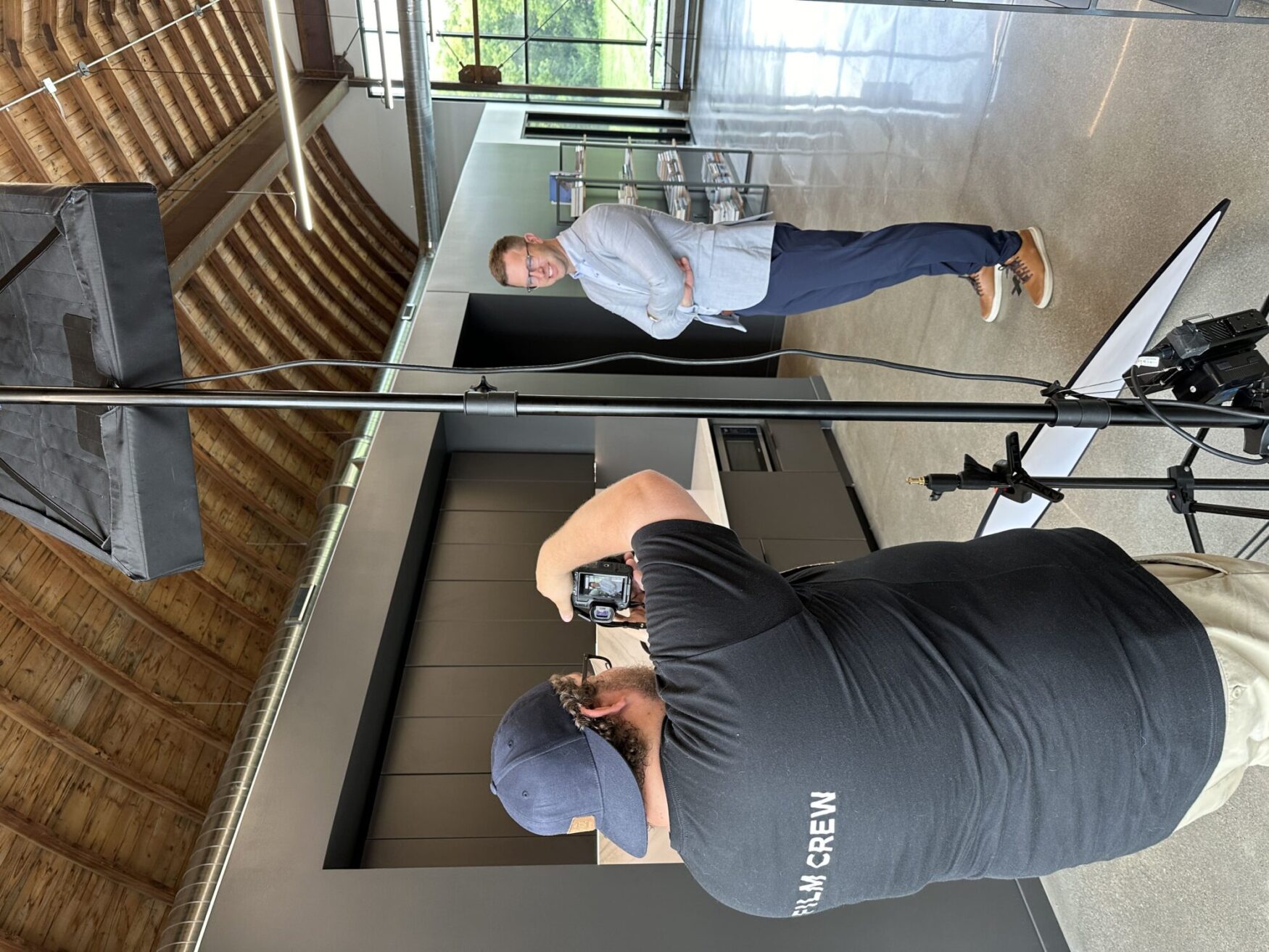 A photographer, wearing a 'FILM CREW' shirt and a cap, is taking a photo of a man standing in an industrial-style building with a curved wooden ceiling. The man being photographed is wearing a light blue blazer, blue pants, and brown shoes, and is smiling with his arms crossed. The setting appears to be a modern office or studio space with large windows and natural light.