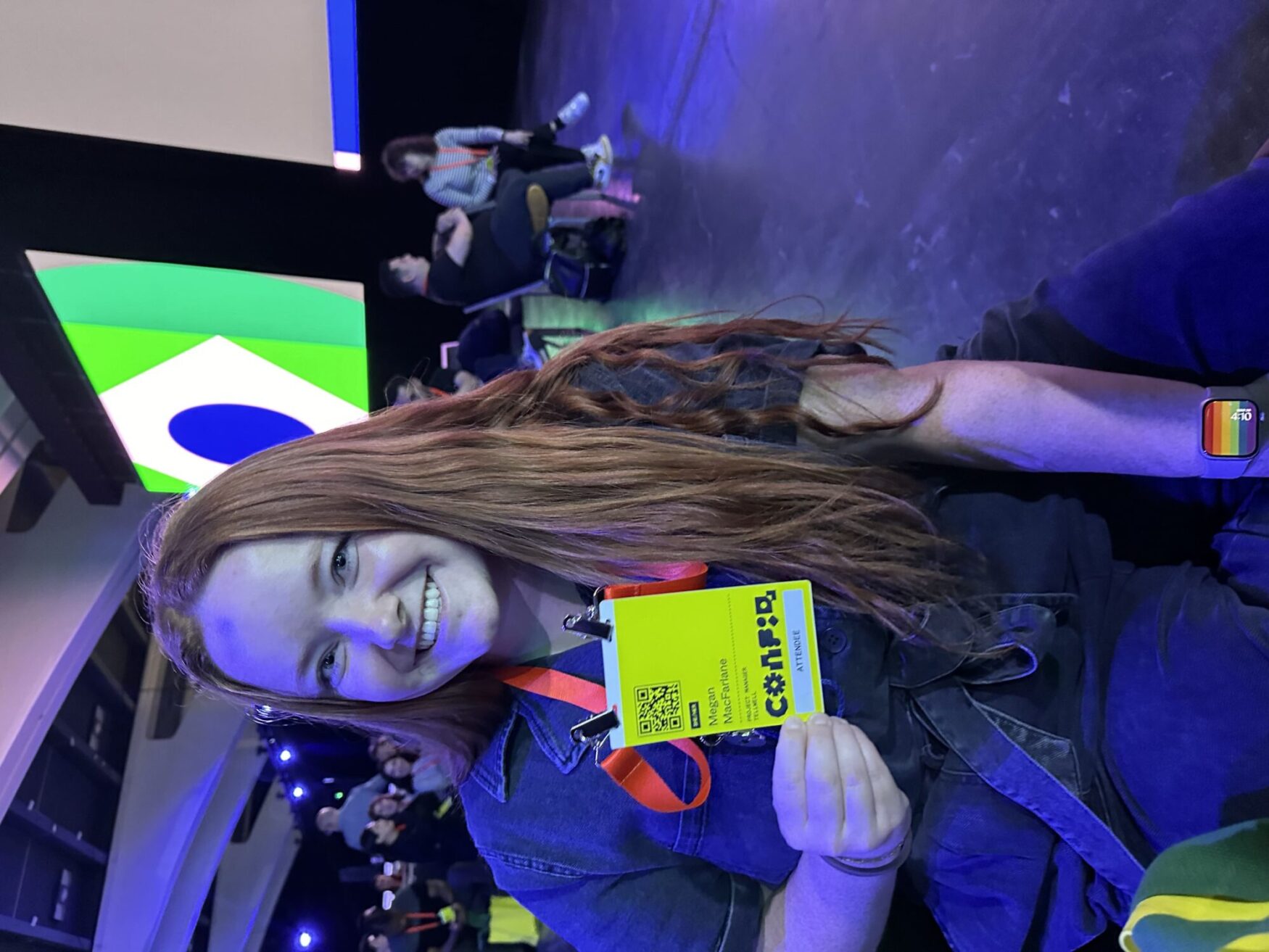 A woman with long red hair is smiling and holding up her conference badge inside a large conference hall. The hall is lit with blue and green lights, and there are large screens displaying colorful graphics in the background. She is wearing a smartwatch with a rainbow Pride band.
