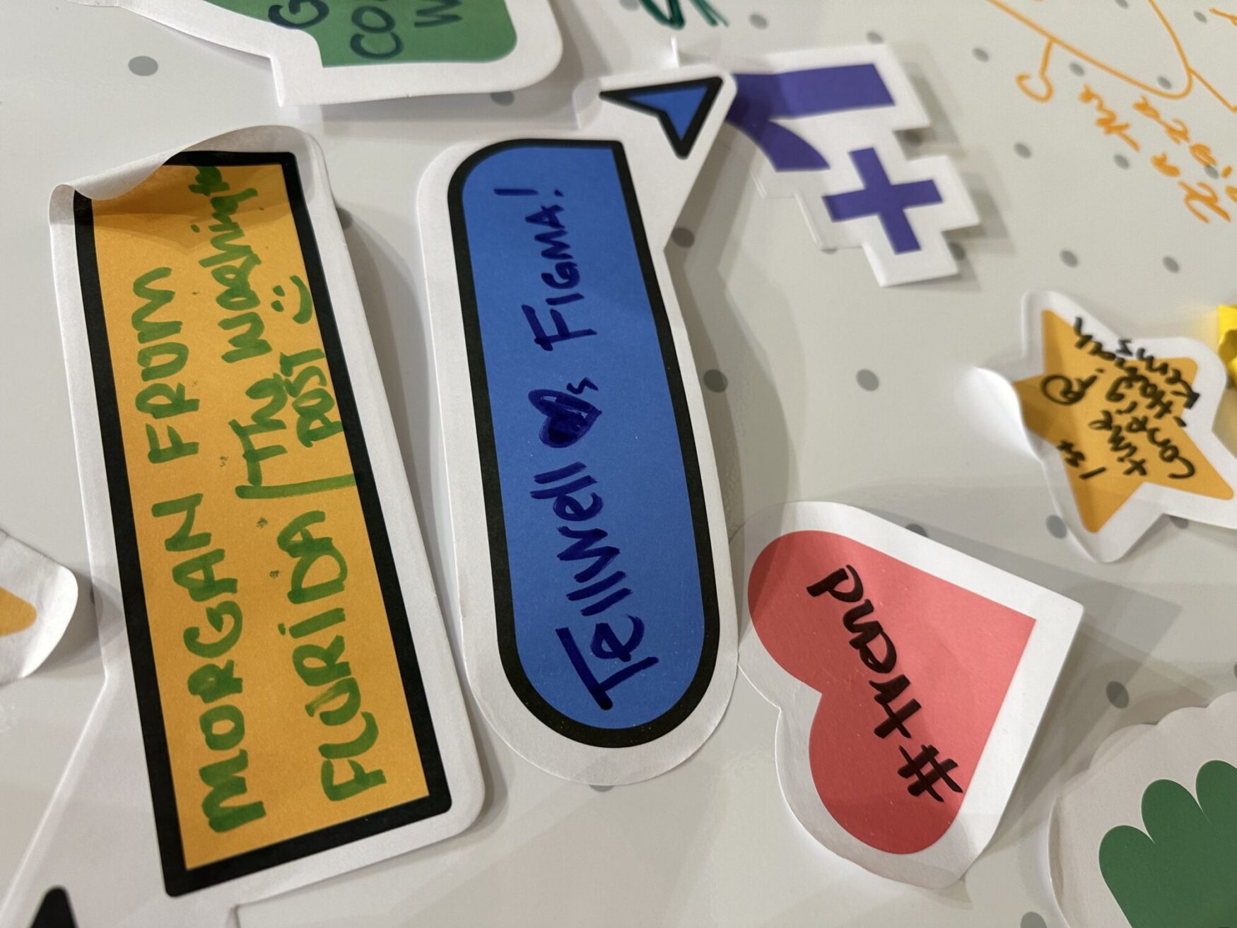 A close-up of various colorful stickers on a wall, with one prominently reading "Tellwell 💜 Figma!" and another from "Morgan from Florida / The Washington Post."