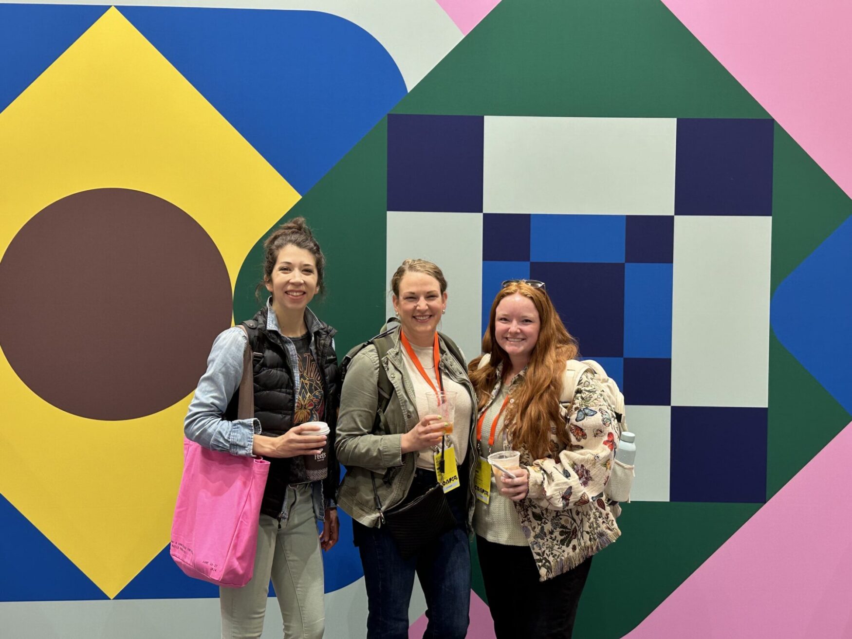 Three women standing in front of a vibrant, geometric mural, smiling and holding drinks, wearing conference badges.