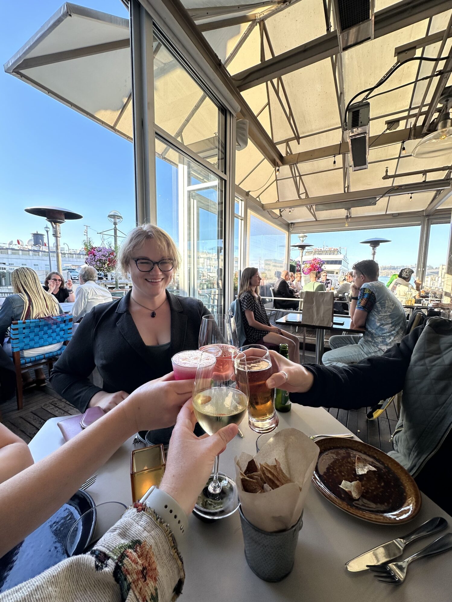A group of people sitting at a restaurant table, toasting with glasses of various drinks. A woman in a black blazer and glasses is smiling in the center.