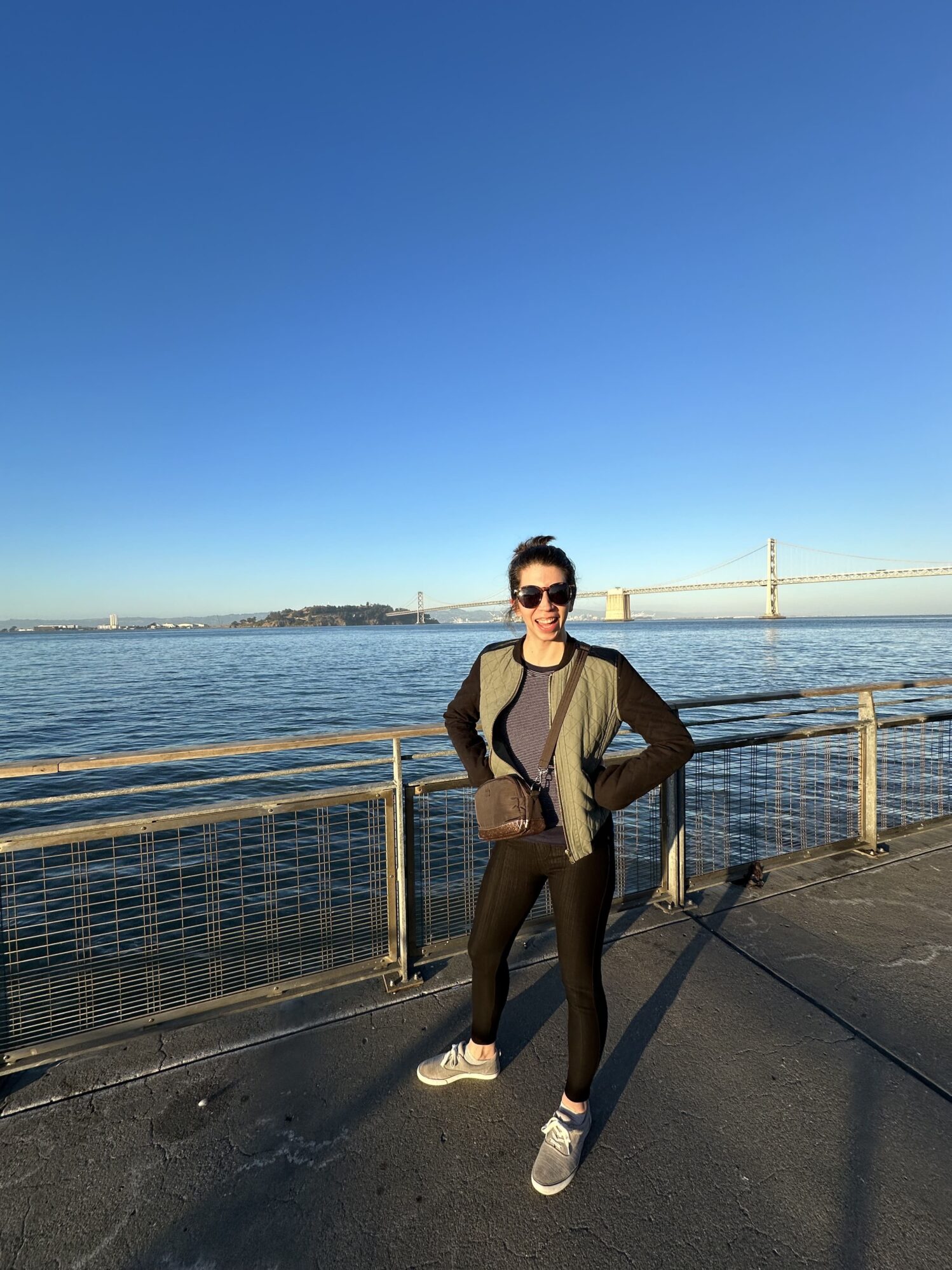 A woman wearing sunglasses and casual clothes poses with hands on her hips by the waterfront, with the San Francisco Bay and a bridge in the background on a sunny day.