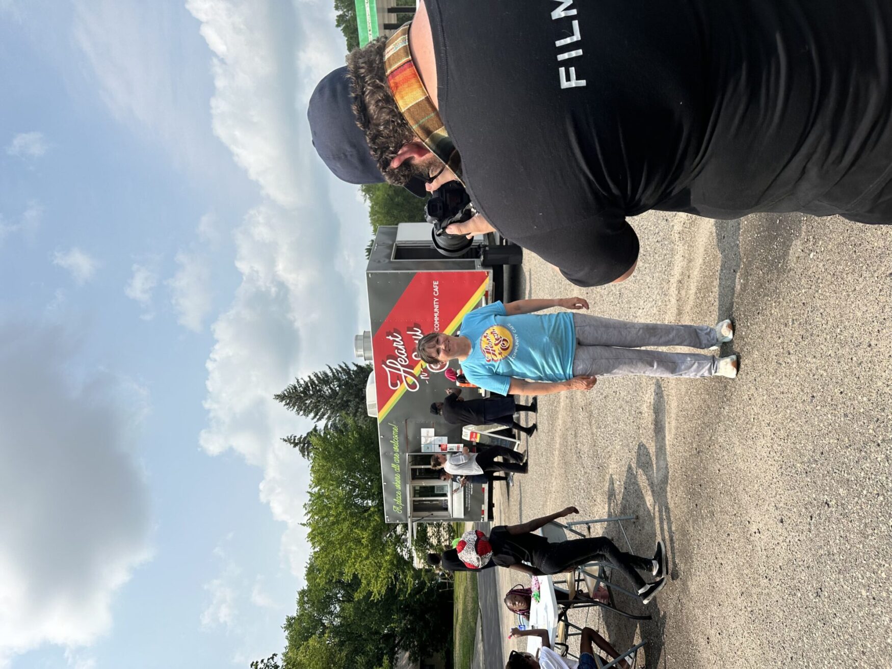 A photographer capturing a photo of a woman standing in front of a food truck with the name 'Heart-n-Soul Community Cafe.' The woman is wearing a light blue 'Heart-n-Soul' T-shirt and light gray pants. In the background, people are gathered around tables and interacting near the food truck. The setting appears to be an outdoor event on a sunny day with a partly cloudy sky and green trees in the background