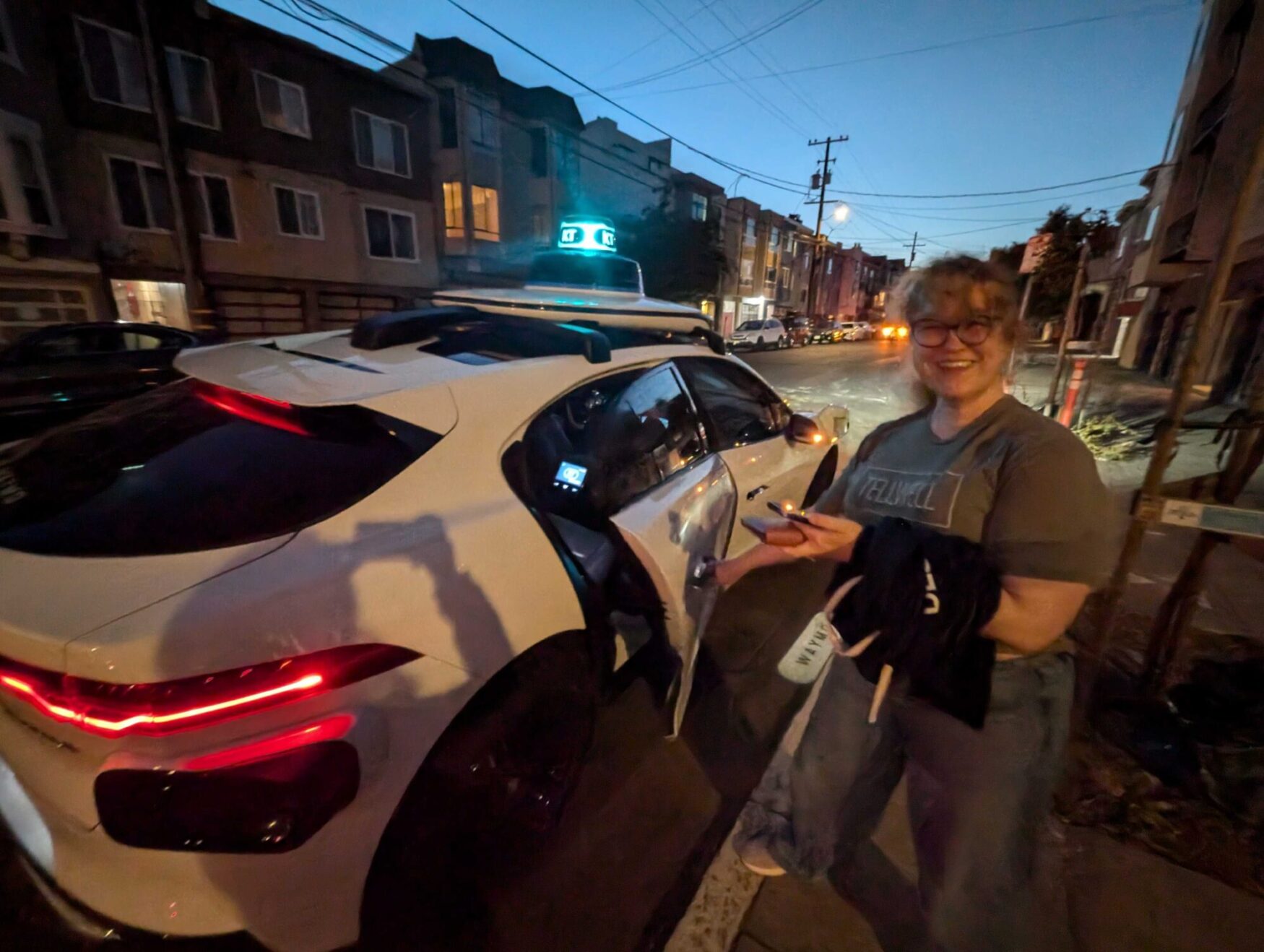 A woman smiling beside a white self-driving car at night, illuminated by street lights.