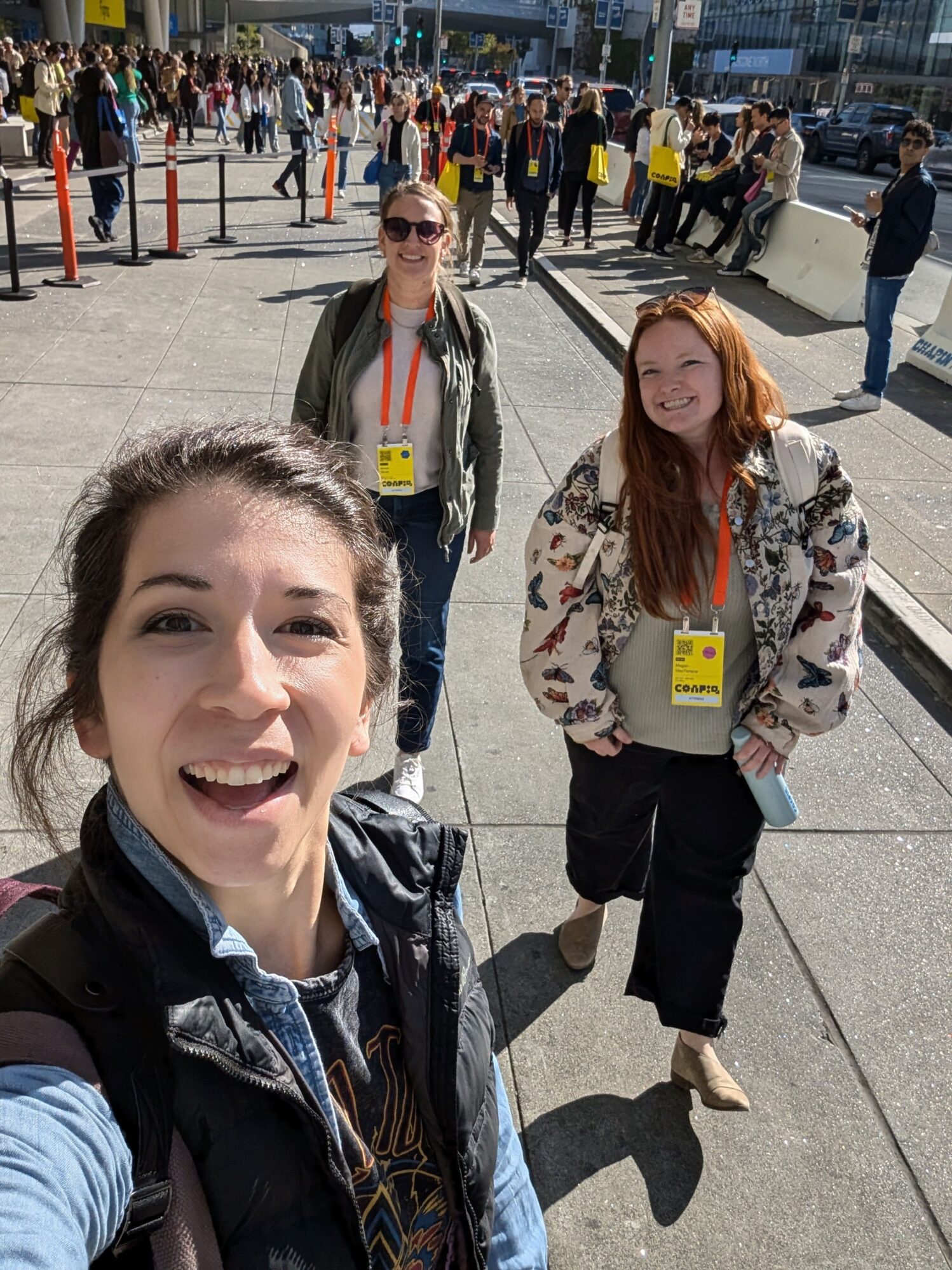 Three women walking down a busy sidewalk at a conference, smiling and wearing name badges with lanyards. One woman is taking a selfie, and others are walking behind her.