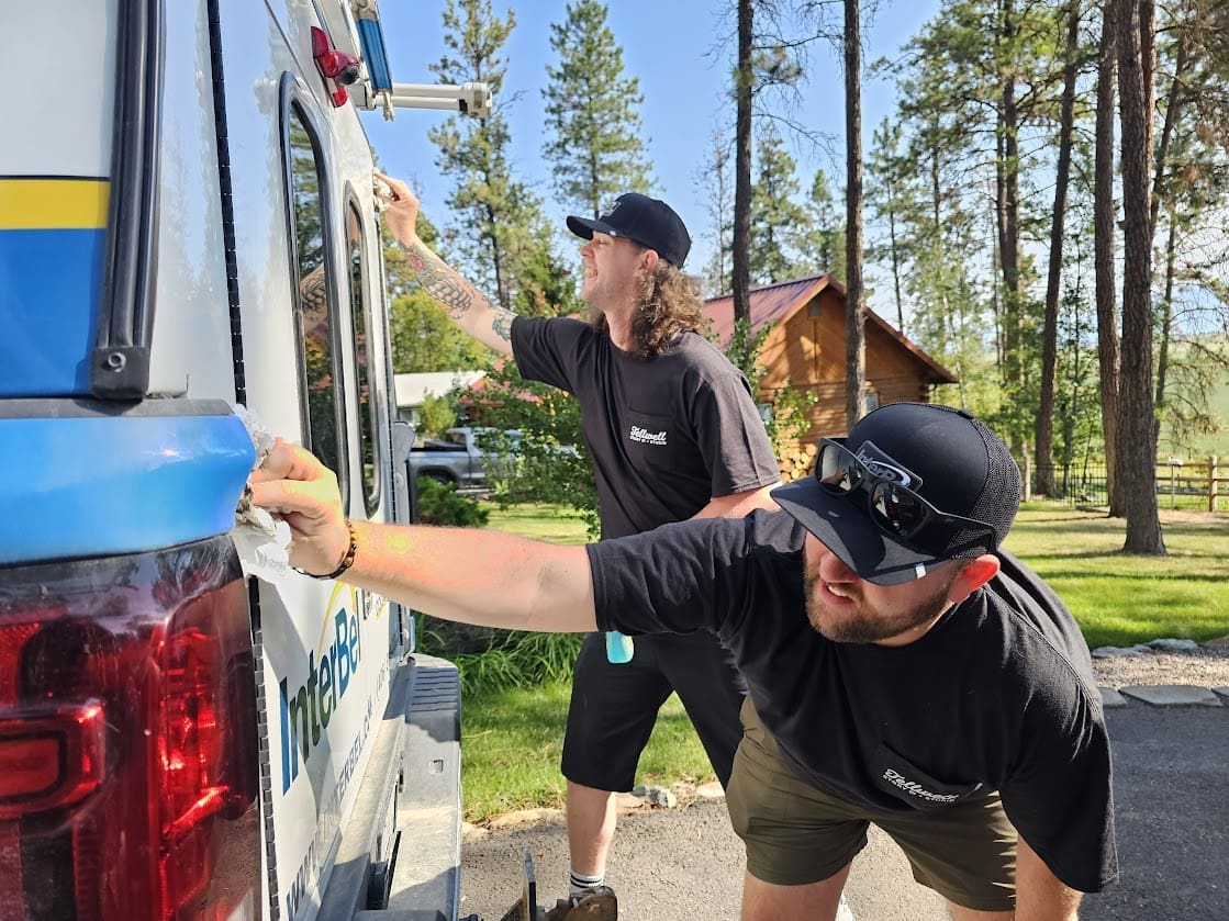 Two people wearing black Tellwell t-shirts and black caps are cleaning a vehicle with white towels. The person on the left, with long curly hair and visible tattoos on their arm, is reaching up to clean a higher spot on the vehicle, while the person on the right is bent over slightly to clean a lower area. The scene takes place outdoors on a sunny day, with trees and a wooden house visible in the background.