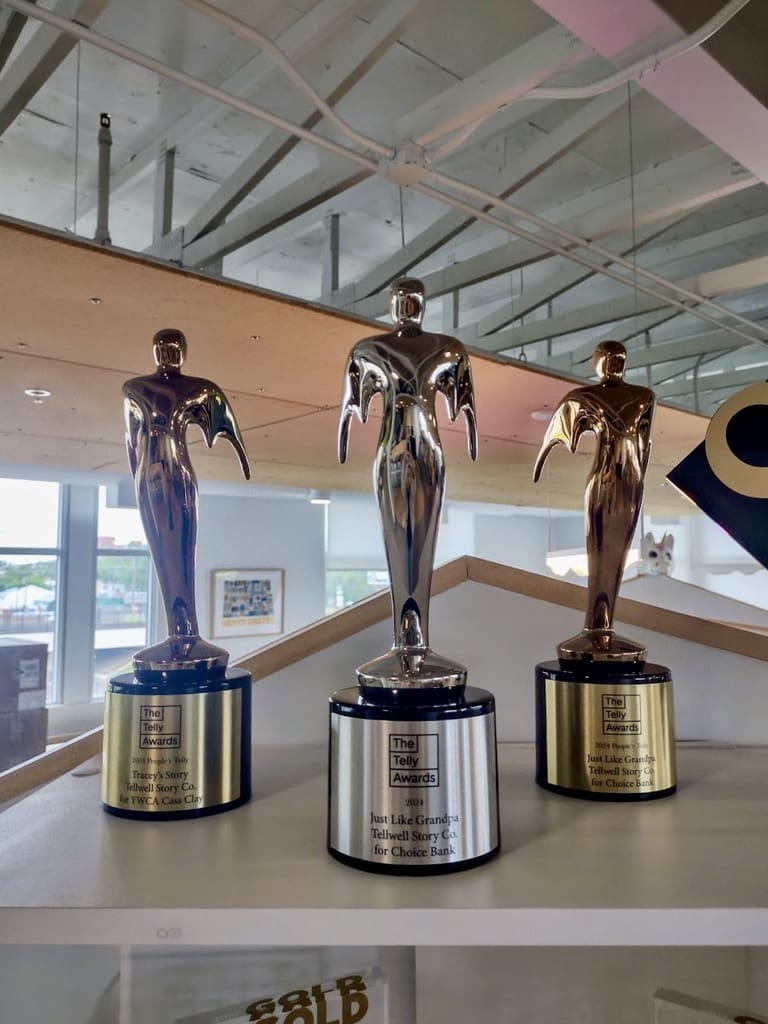A close-up image of three Telly Awards trophies displayed on a white shelf. The trophies are shiny, metallic statuettes with wings and stand on round bases with engraved plaques. The plaques on two trophies read "2024 People's Telly - Tracey's Story - Tellwell Story Co. for YWCA Cass Clay" and "2024 Bronze Telly - Just Like Grandpa - Tellwell Story Co. for Choice Bank." The middle trophy, which has a silver finish, reads "2024 Silver Telly - Just Like Grandpa - Tellwell Story Co. for Choice Bank." The background shows a modern office setting with large windows and exposed ceiling beams.