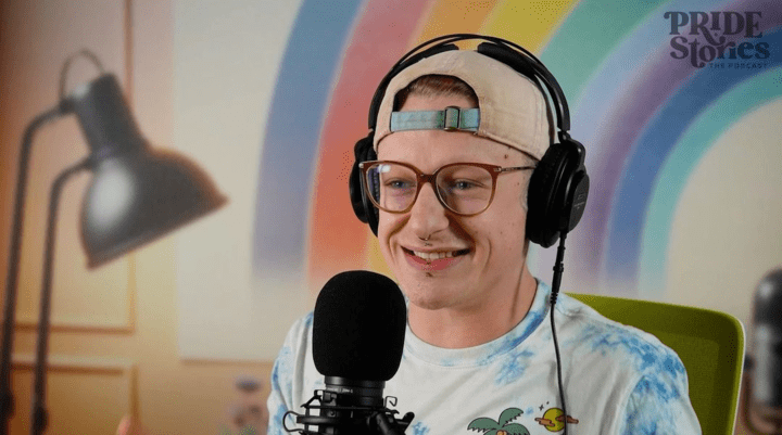 A person wearing a backward baseball cap, glasses, and a tie-dye shirt is sitting in front of a microphone, recording for a podcast. They are smiling and wearing large over-ear headphones. In the background, there is a colorful rainbow mural and a blurred-out desk lamp. The podcast logo, "PRIDE Stories," is visible in the upper right corner of the image. The setting is bright and vibrant, giving off a cheerful and welcoming vibe