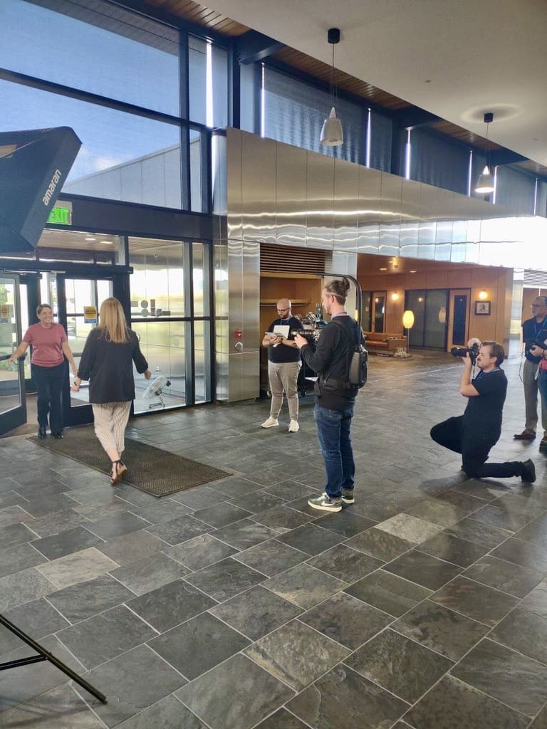 A group of people are filming a scene in a spacious, modern building with large glass windows and stone tile floors. One woman in a black jacket and striped pants is walking toward another woman in a red shirt near the entrance, which has glass doors. The crew includes a person kneeling with a DSLR camera, another standing with a video camera, and a third person holding a clipboard. A large light is set up on the left side of the image, indicating a professional video shoot in progress. The atmosphere is focused and professional.