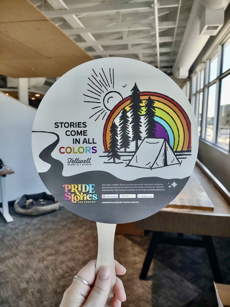 A hand is holding up a circular paper fan with a wooden handle. The fan features an illustration of a campsite with trees, a tent, and a sun in the background. A rainbow arcs over the scene, adding vibrant colors to the design. The text on the fan reads, "STORIES COME IN ALL COLORS," followed by "Tellwell Story Co. + Studio" and "PRIDE Stories - The Podcast." The bottom of the fan includes logos for Apple Podcasts, Spotify, and Google Podcasts, along with a web address for the Pride Stories podcast. The background shows a modern office space with exposed ceilings and large windows.