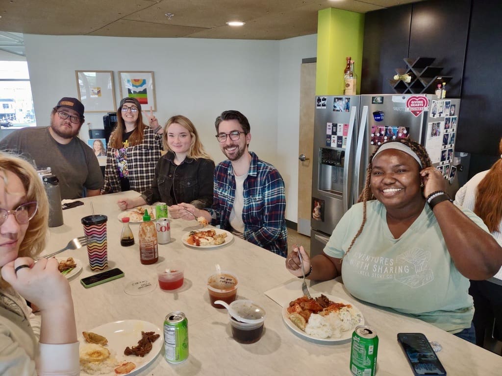 A group of people are sitting around a table in a kitchen area, enjoying a meal together. They are smiling and chatting, with plates of food and drinks in front of them. The atmosphere is casual and friendly, with a mix of food condiments and drinks on the table, including sriracha, soy sauce, and cans of soda. Behind them, there is a refrigerator covered with photos and magnets, and a small wine rack. The scene conveys a sense of community and camaraderie as they share a meal.