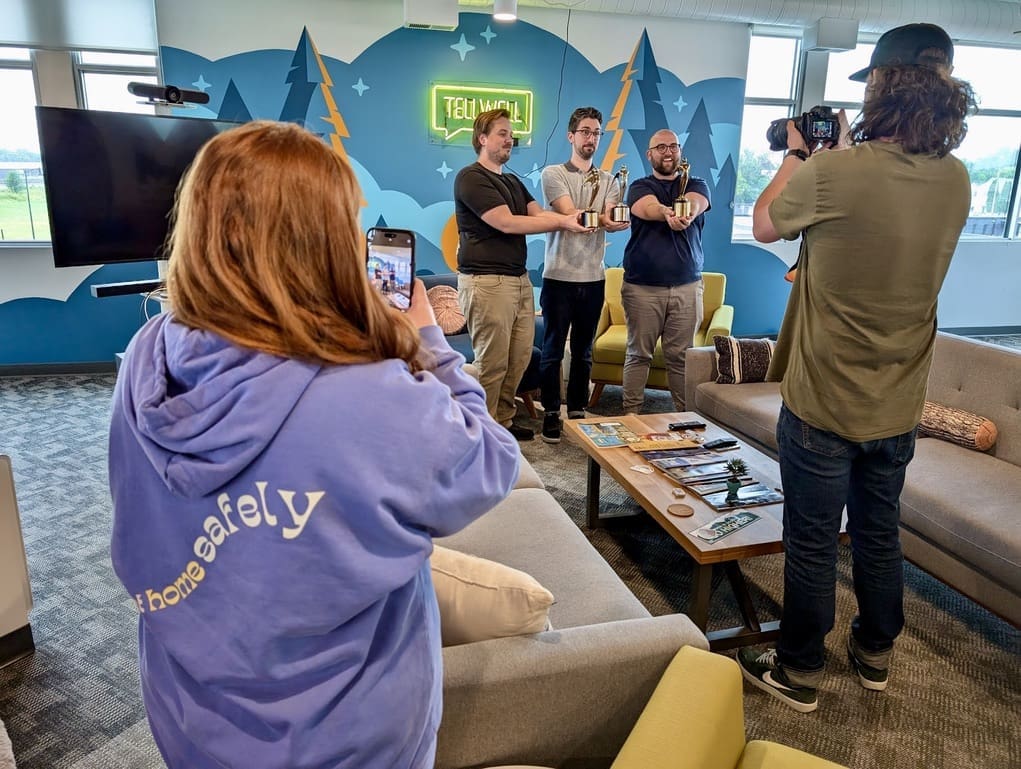 In an office setting with colorful wall art and a neon "TELLWELL" sign, three men stand side by side, smiling and holding up gold statuette awards. They are being photographed by a person with long hair, wearing a baseball cap, who is holding a DSLR camera. Another person, wearing a light purple hoodie with the words "Get home safely" on the back, is taking a photo of the group with a smartphone. The room has a casual and creative atmosphere, with comfortable seating, a coffee table with various items, and large windows letting in natural light.