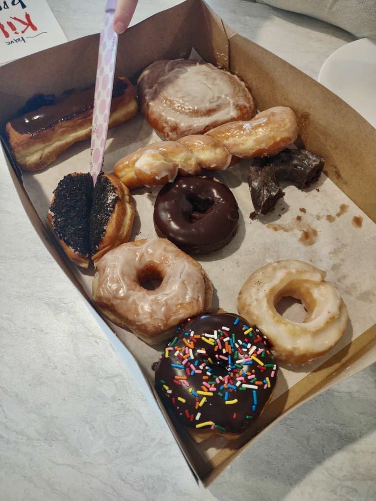 A close-up of an open box of assorted donuts, including a chocolate-frosted donut with sprinkles, glazed donuts, a twisted glazed pastry, a chocolate-frosted eclair, and other varieties. A hand holding a pink and white polka-dotted knife is cutting one of the donuts in half. Part of a sign that reads 'Killer birthday' is visible in the upper left corner of the image.