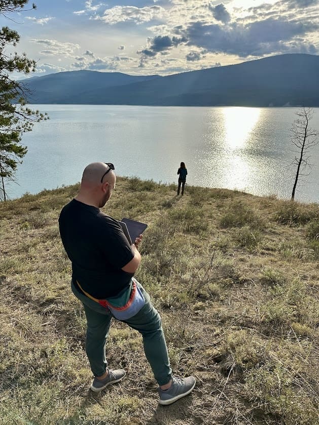 A bald person wearing a black shirt and green pants is holding a tablet while standing on a grassy hill overlooking a large, calm lake. The person is facing away from the camera, and another person is standing near the water's edge in the distance. The sun is low in the sky, casting a bright reflection on the lake, and the surrounding mountains create a serene backdrop.