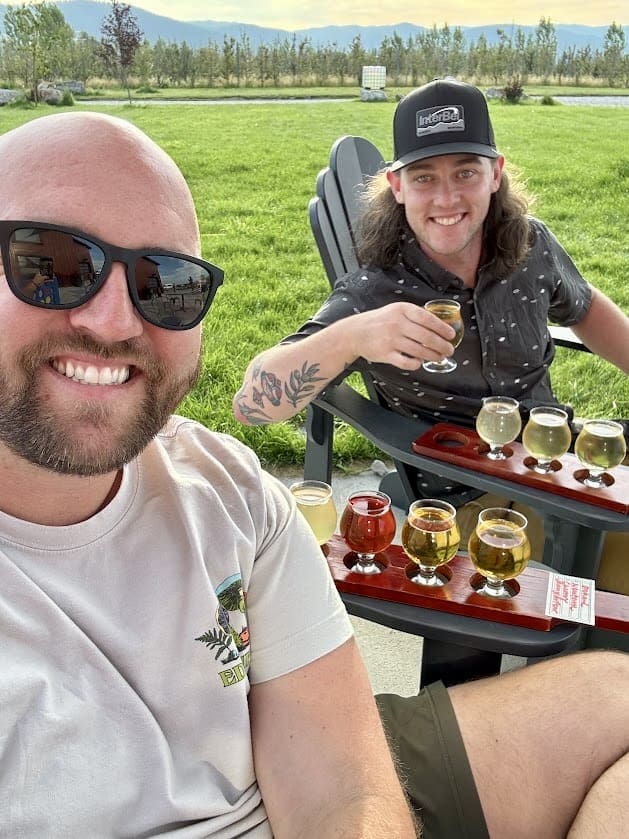 Two people are sitting outdoors on a sunny day, smiling at the camera while enjoying a flight of beer. The person on the left is bald, wearing sunglasses, and a light-colored t-shirt, while the person on the right has long curly hair, is wearing a black hat, and a dark patterned shirt. Both have trays of various beer samples in front of them. They are seated in black outdoor chairs on a grassy area with a scenic background of trees and mountains