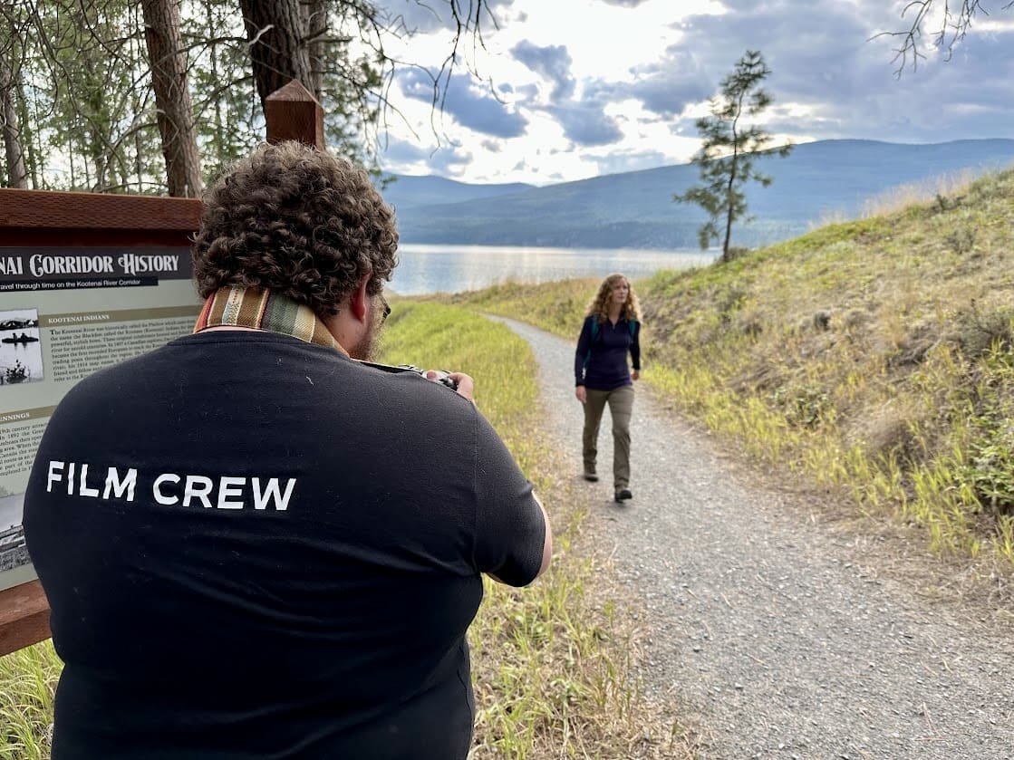 A person wearing a black shirt with the words 'FILM CREW' on the back is taking a photo or filming another person walking along a gravel path near a lake. The path is surrounded by tall grass, and the lake is bordered by mountains in the distance. The sky is partly cloudy, creating a scenic outdoor setting. The person being filmed is dressed casually in outdoor clothing, and a sign with historical information about the Kootenai Corridor is visible in the background.