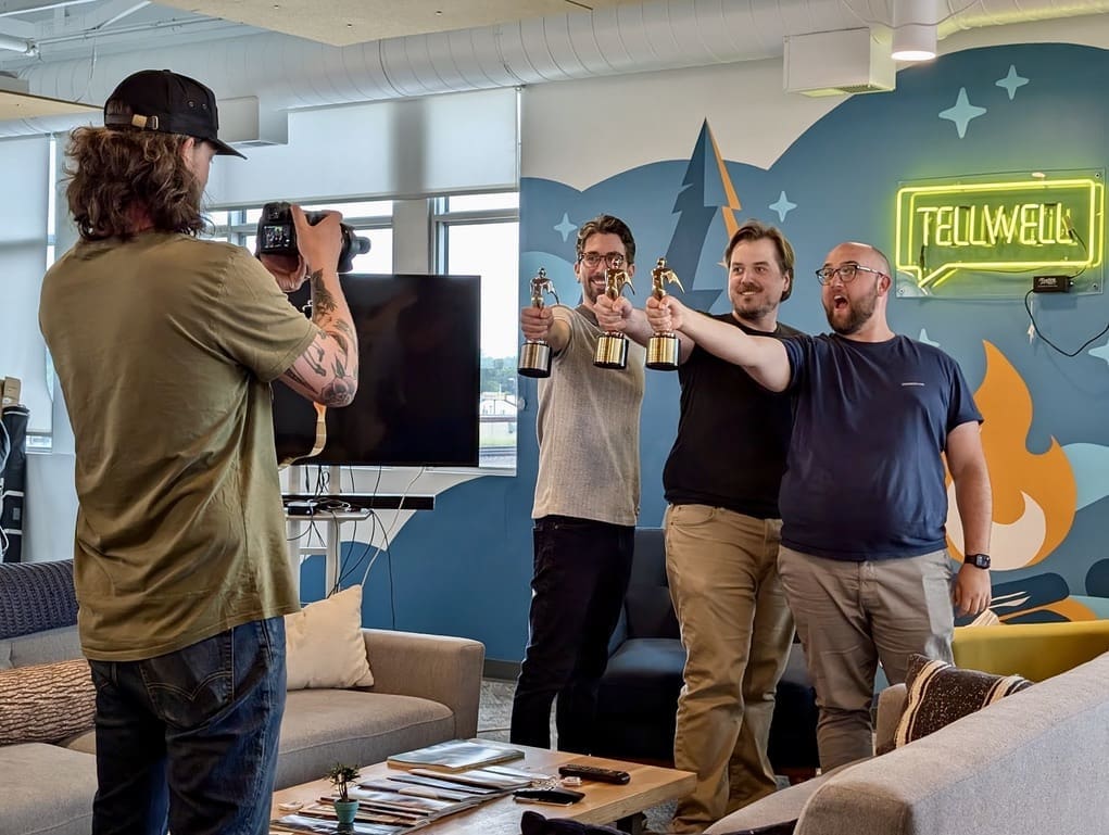 A person with long hair, a beard, and a baseball cap is taking a photo with a DSLR camera. They are photographing three men who are standing side by side, smiling, and holding up gold awards shaped like statuettes. The men appear to be in an office with modern decor, including a neon sign that reads "TELLWELL" on the wall behind them. The atmosphere is casual and celebratory.