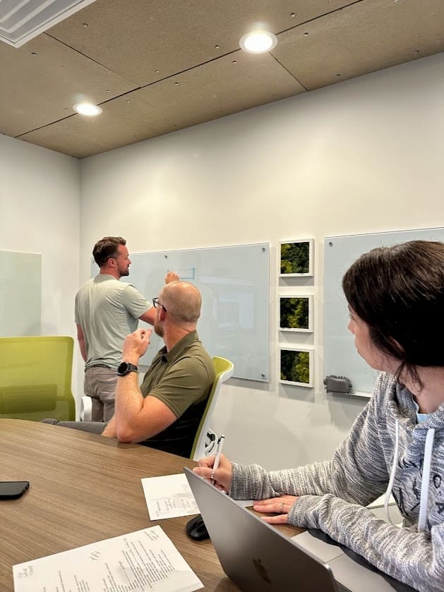 Three people are in a meeting room. One person is standing at the front, writing on a whiteboard, while the other two are seated at a table, facing the whiteboard. The person standing is wearing a light green shirt, and the person closest to the camera is taking notes on a laptop while wearing a gray hoodie. The third person, who is bald, is seated in the middle, attentively watching the whiteboard. Papers and a laptop are spread out on the table in front of them.
