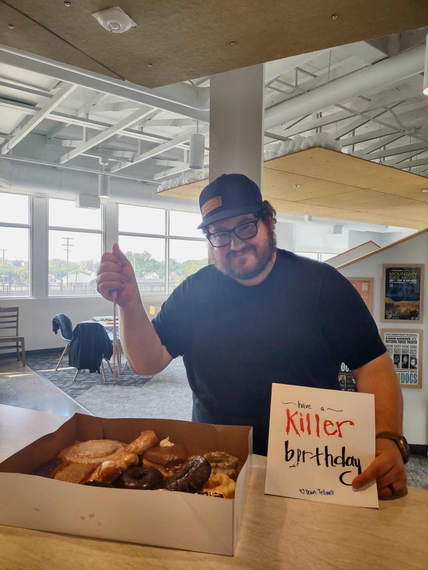 A person with curly hair and glasses, wearing a black t-shirt and a dark cap with a brown patch, is standing in an office space holding a large box of assorted donuts. They are smiling and giving a thumbs-up with one hand while holding a sign in the other hand that reads, 'Have a killer birthday - Team Tellwell!' The office space behind them has large windows and modern decor.