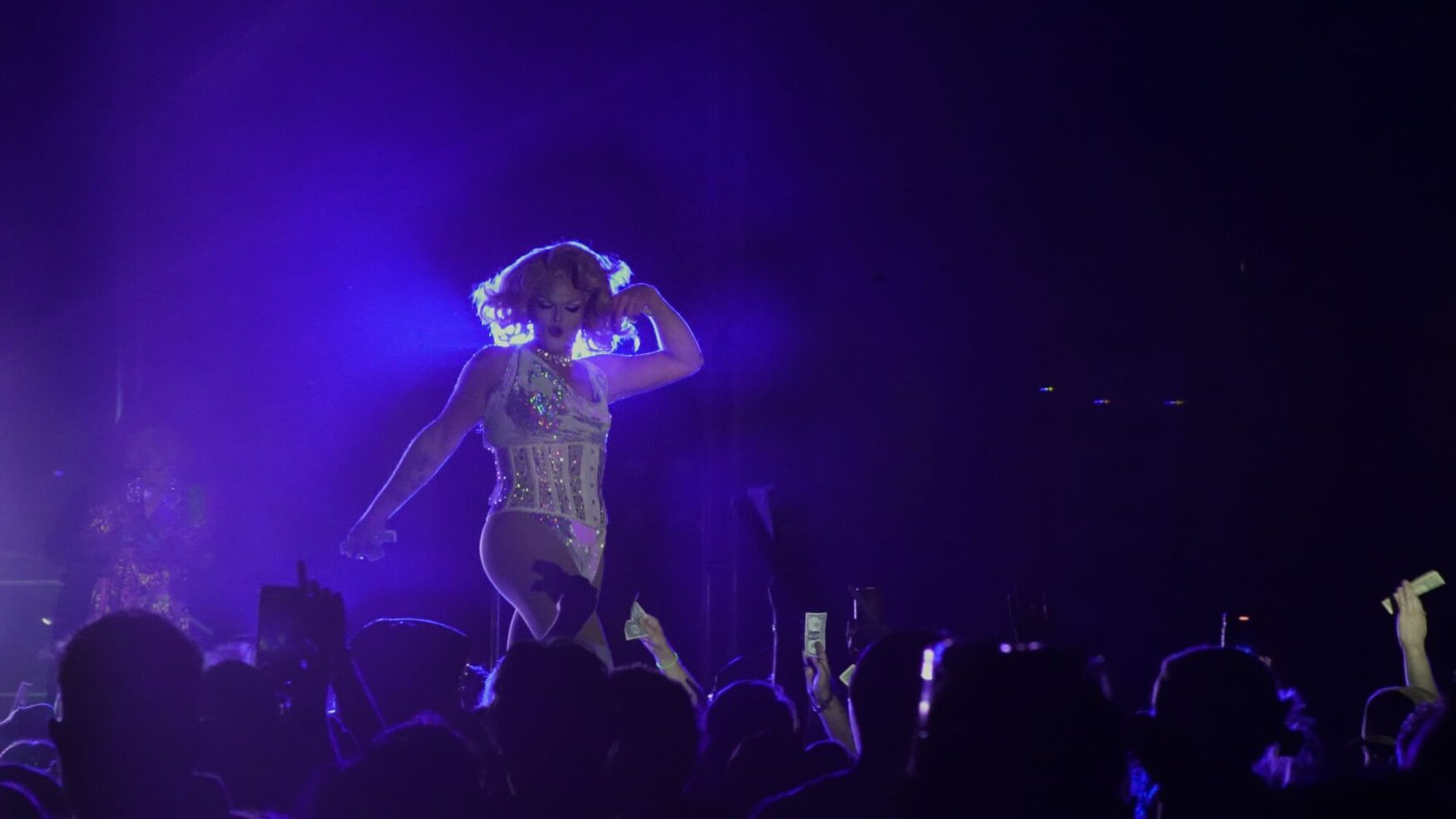 A drag queen struts her stuff, backlit with purple light on a stage