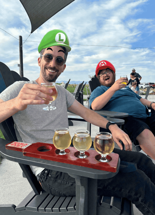 Two people are sitting outdoors in black Adirondack chairs, smiling and holding up glasses of beer. The person on the left is wearing sunglasses, a gray t-shirt, and has short hair with a bit of gray, while the person on the right is wearing a cap and a blue t-shirt. Both have trays with multiple beer samples in front of them. The scene takes place on a sunny day with a blue sky and light clouds, and the background includes other people and a view of the outdoors