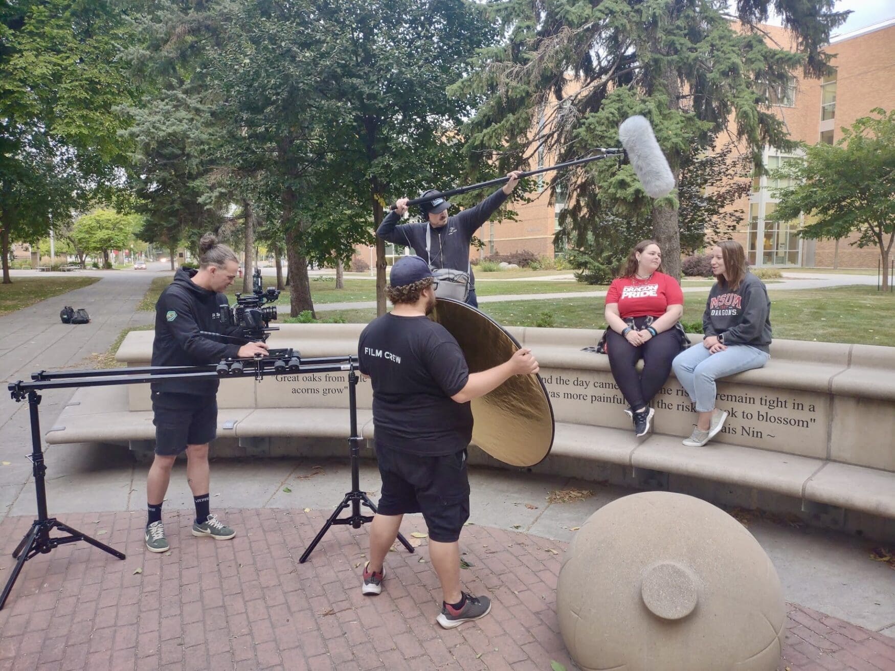 Film crew setting up camera, microphone, and lighting equipment while two women sit on a stone bench talking on a university campus. Trees and a building are visible in the background.