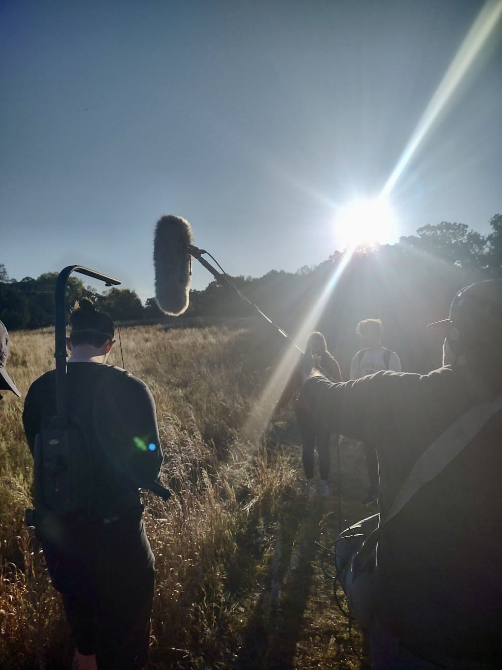 A film crew is outdoors in a grassy field, walking towards the sun as it sets or rises in the sky. One person holds a boom microphone, while others walk ahead, silhouetted by the bright sunlight. The scene is bathed in natural light, creating long shadows and a cinematic atmosphere.
