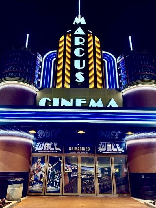 The illuminated entrance of a Marcus Cinema at night, featuring an art-deco style marquee with bold vertical lines and neon lights. The large, vertical sign reads 'MARCUS' above the word 'CINEMA.' Below the marquee, the doors are adorned with movie posters and advertisements, including one for 'The Wall Reimagined.'