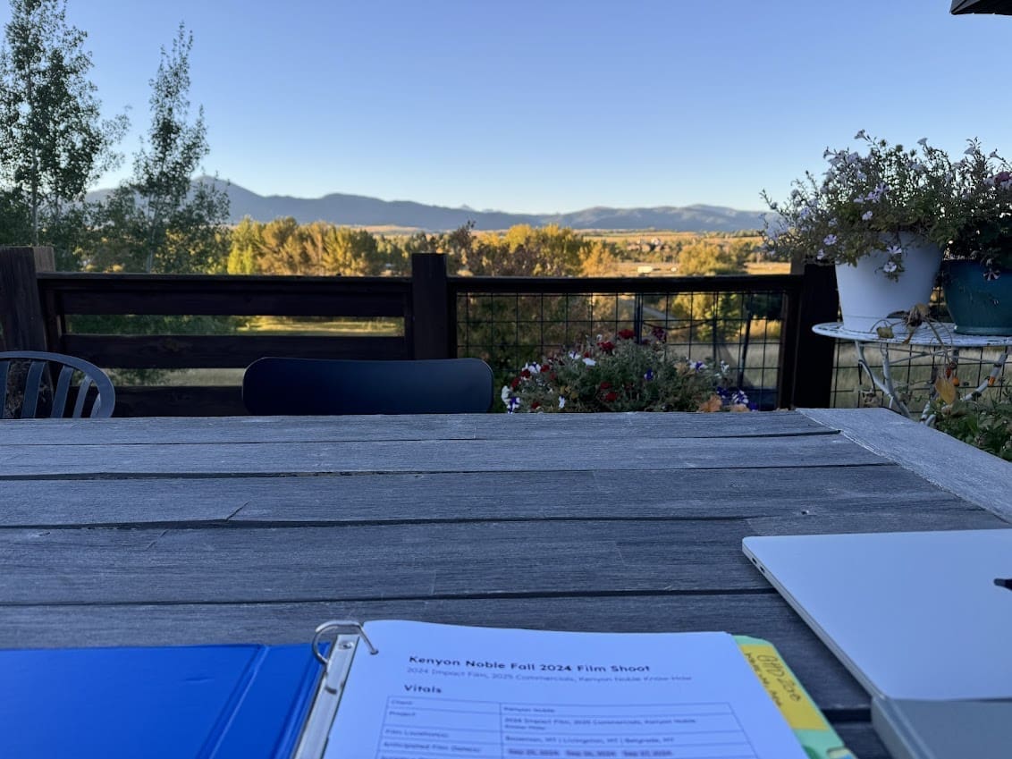 An outdoor patio with a wooden table displaying a binder titled 'Kenyon Noble Fall 2024 Film Shoot' and a laptop. In the background, trees, fields, and mountains are visible under a clear blue sky.