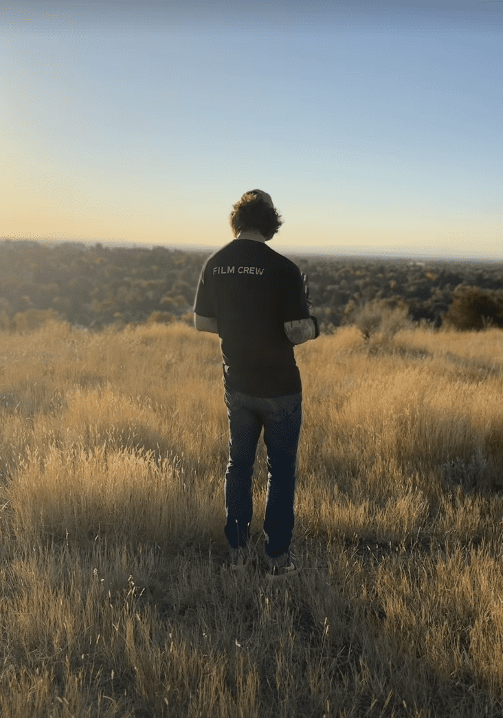 A person wearing a black 'FILM CREW' shirt stands in a field of tall, golden grass, holding a camera while looking out toward a distant tree line during sunrise or sunset.