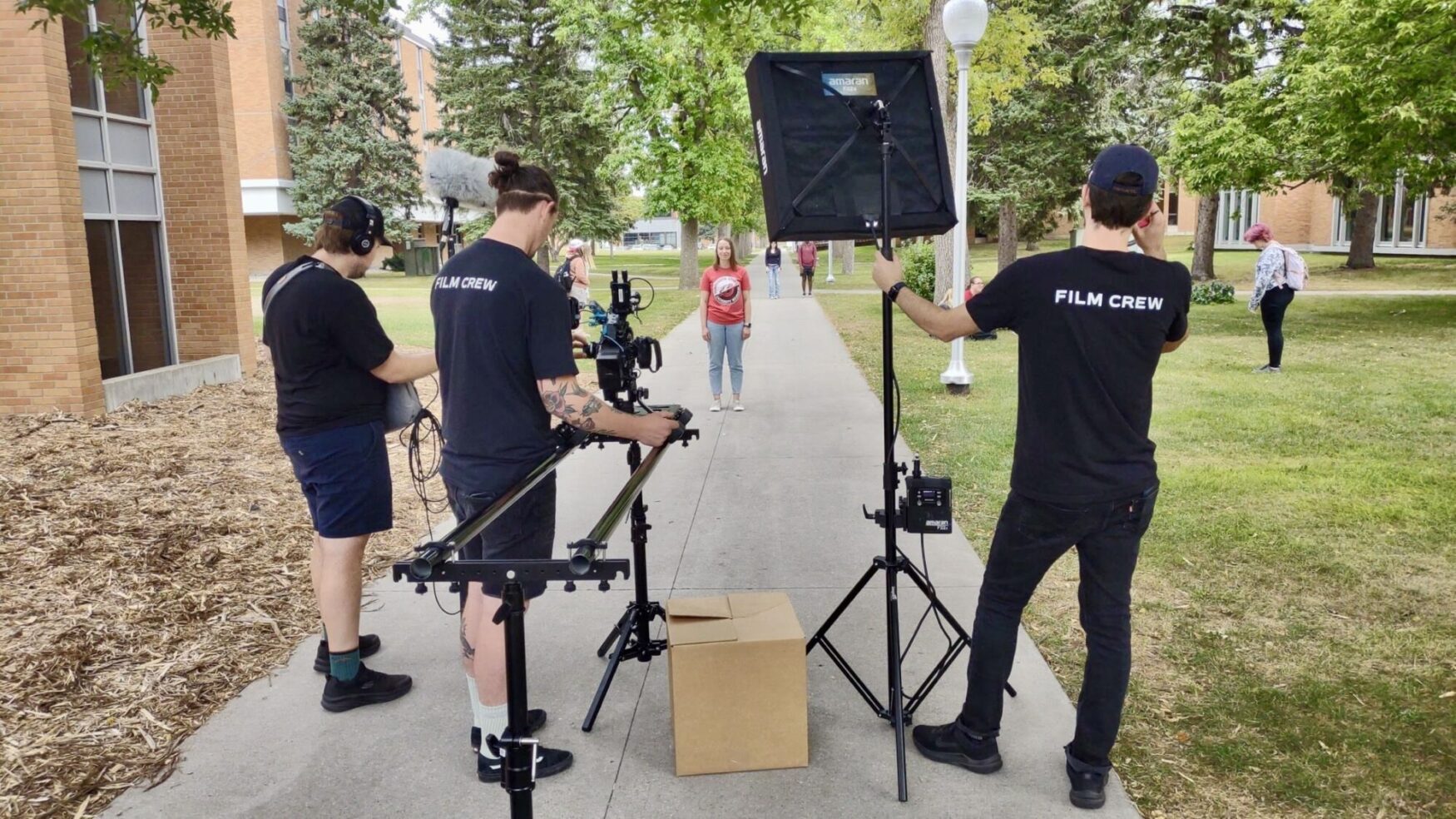 A film crew is set up outdoors on a college campus, filming a woman standing on a pathway. One crew member operates a camera on a slider, another holds a boom microphone, and a third adjusts a large light. All crew members are wearing 'Film Crew' shirts. The setting features brick buildings, trees, and a grassy area with other people walking in the background.